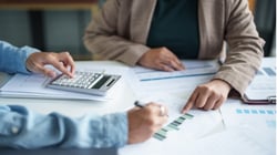Two people reviewing financial documents at a desk, pointing to charts and using a calculator to analyze data together.