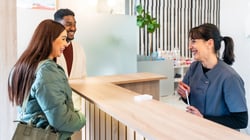 A dental receptionist in blue scrubs smiles and speaks with two patients at a modern reception desk.