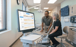 A dentist and patient reviewing a digital periodontal chart on a computer monitor inside a modern dental treatment room.