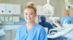 A smiling dental assistant in blue scrubs standing in a dental operatory, with a colleague and dental chair visible in the background.