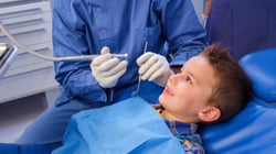 A young boy smiling up at a dentist during a pediatric dental exam, with the dentist holding a dental drill and mirror