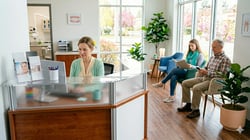 Dental front office administrator working at a modern reception desk in a bright, clean dental practice with patients waiting in the background.