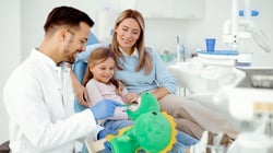 Pediatric dentist showing a young girl and her mother how to brush teeth using a dinosaur toy during a dental visit.
