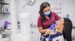 A dental hygienist in maroon scrubs and a blue face mask performing a dental procedure on a female patient reclined in a dental chair in a modern dental office.