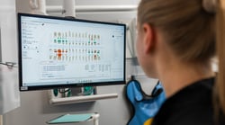 A dental hygienist with blonde hair pulled back, wearing gloves, views a patient's perio chart and tooth diagram displayed in Curve Dental's cloud-based practice management software on a wall-mounted ASUS monitor in a dental treatment room.