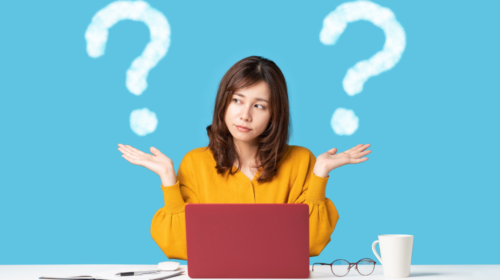 Woman in a yellow sweater sitting at a desk with a red laptop, shrugging with question marks behind her on a blue background, with glasses and a coffee mug nearby.