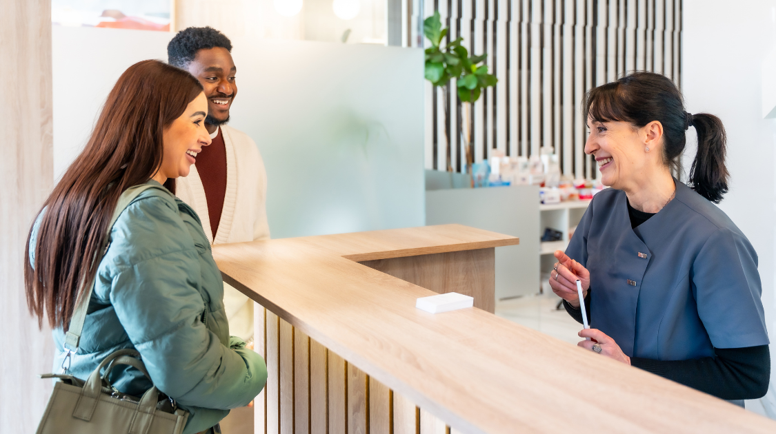 A dental receptionist in blue scrubs smiles and speaks with two patients at a modern reception desk.