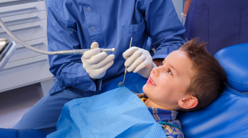 A young boy smiling up at a dentist during a pediatric dental exam, with the dentist holding a dental drill and mirror
