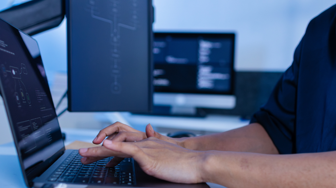 A person typing on a laptop at a multi-monitor workstation displaying code and software interfaces in a dimly lit, blue-toned workspace.