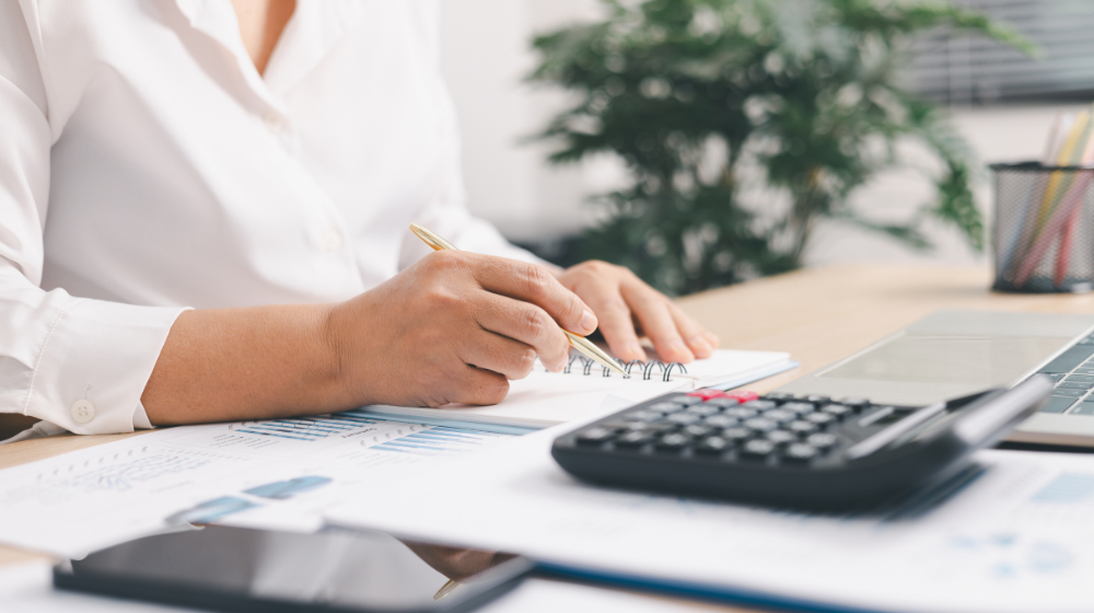 A person in a white shirt writing in a spiral notebook at a desk with financial charts, a calculator, and a laptop.