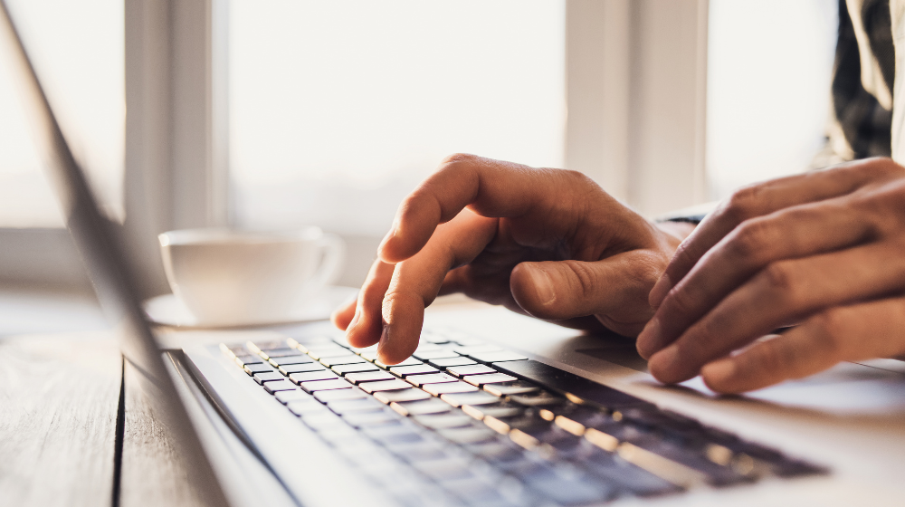 A person typing on a laptop with a coffee cup in the background in a well-lit office.