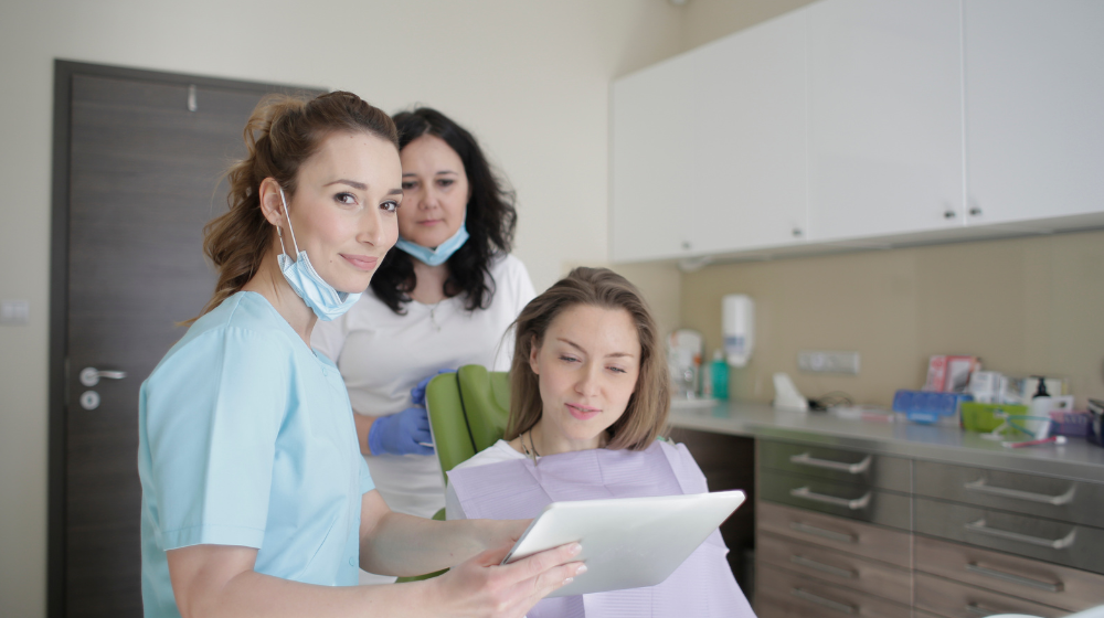 A dental hygienist in teal scrubs shows a tablet to a seated patient in a dental chair, while a second dental professional in a white coat looks on in the background of a modern dental office.