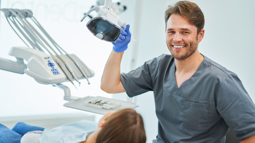Dentist smiling with his left hand on overhead light. Patient in chair looking at dentist.