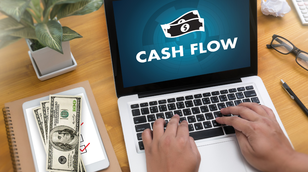 Person typing on a laptop with the words “Cash Flow” and a money icon on the screen, symbolizing financial management and business revenue. Nearby are dollar bills, a notebook, glasses, and a plant on a wooden desk.